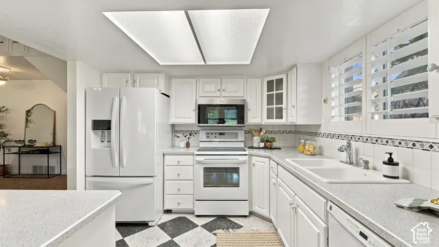White cabinetry with matte silver hardware, white range with overhead microwave, dual basin sink, white refrigerator, and black and white checkered floor tile