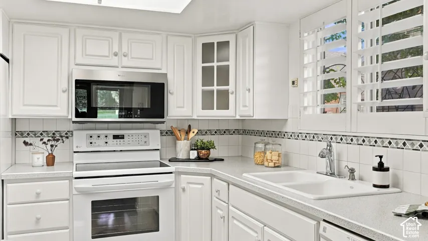 White cabinetry, stainless steel microwave, white electric range, white tile backsplash with decorative accent, and white double basin sink