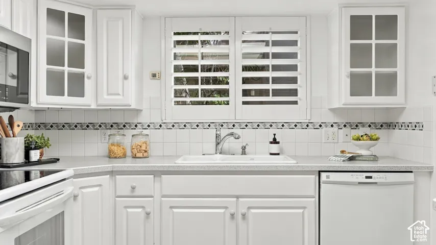 White cabinetry with glass-front accents, white tile backsplash with decorative border, built-in dishwasher, white sink with chrome faucet, and plantation shutters