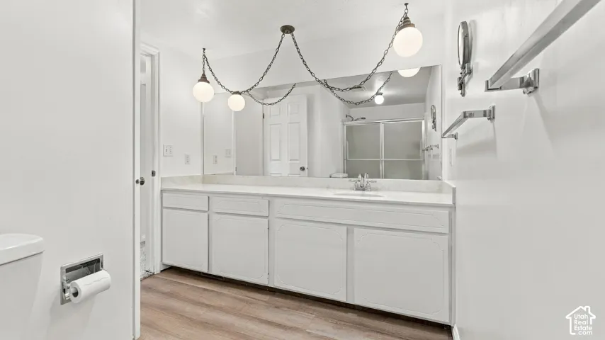 Dual pendant light fixtures, wood-finish flooring, white vanity with integrated sink, large wall-mounted mirror, and a shower with frosted glass enclosure
