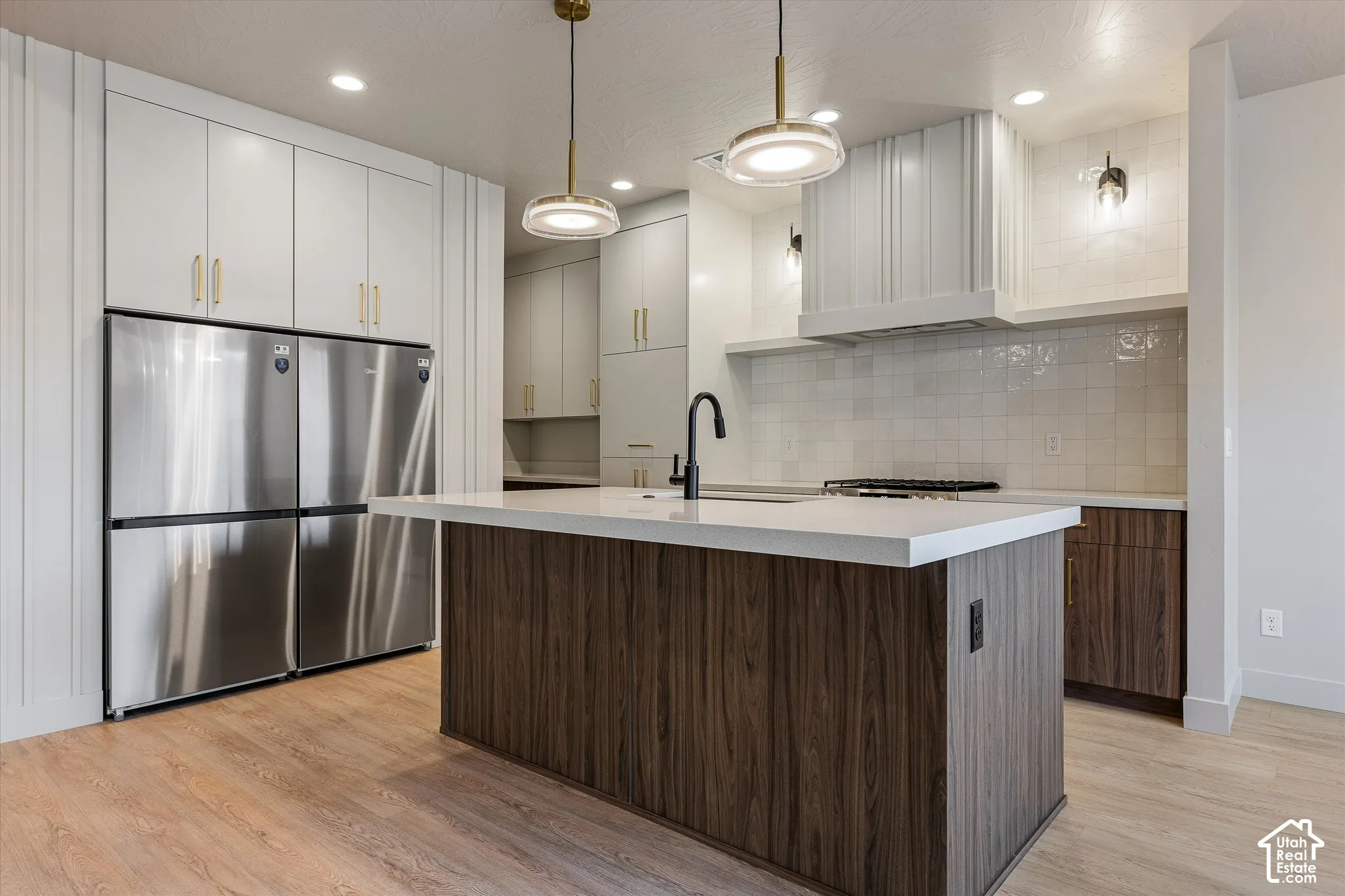 Modern kitchen featuring an expansive center island with a light-toned countertop, stainless steel refrigerator, white upper cabinetry with gold-toned hardware, and a white tiled backsplash