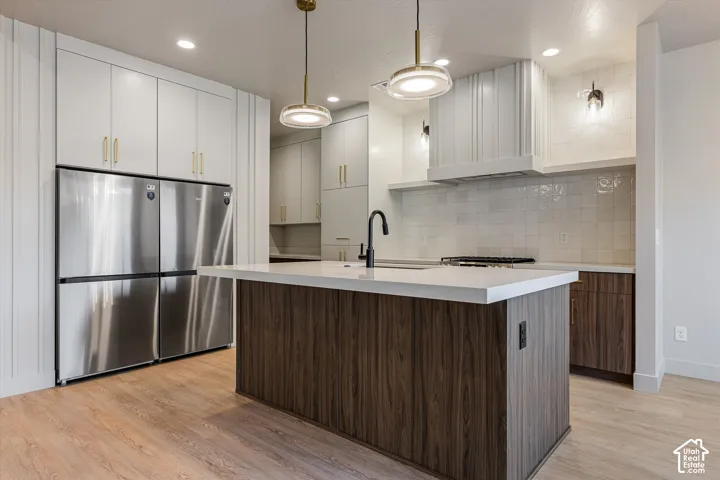 Modern kitchen featuring an expansive center island with a light-toned countertop, stainless steel refrigerator, white upper cabinetry with gold-toned hardware, and a white tiled backsplash