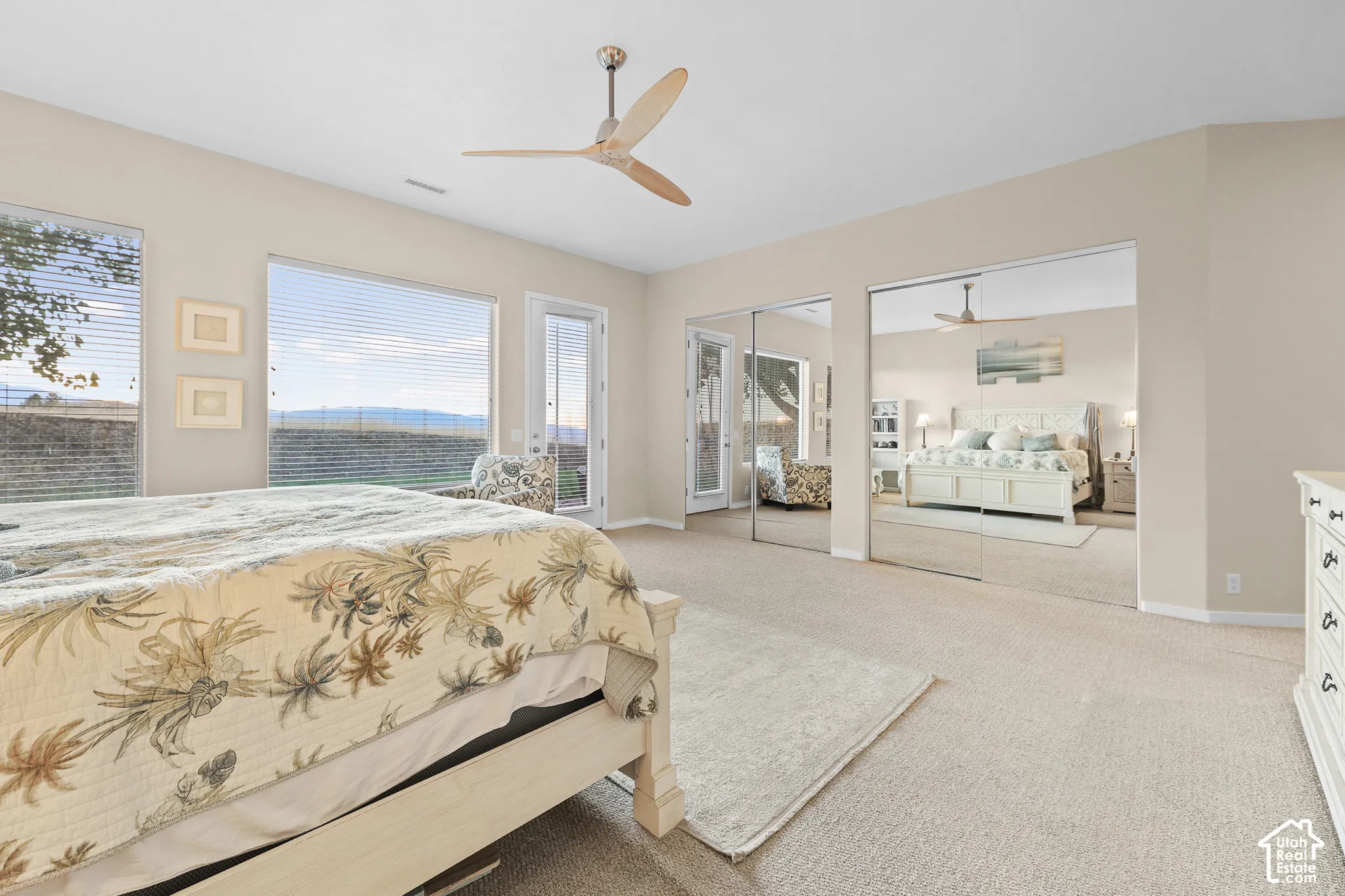 Recessed lighting and a ceiling fan with wood-finish blades illuminate a carpeted room featuring a large window, a glass door, and mirrored sliding closet doors