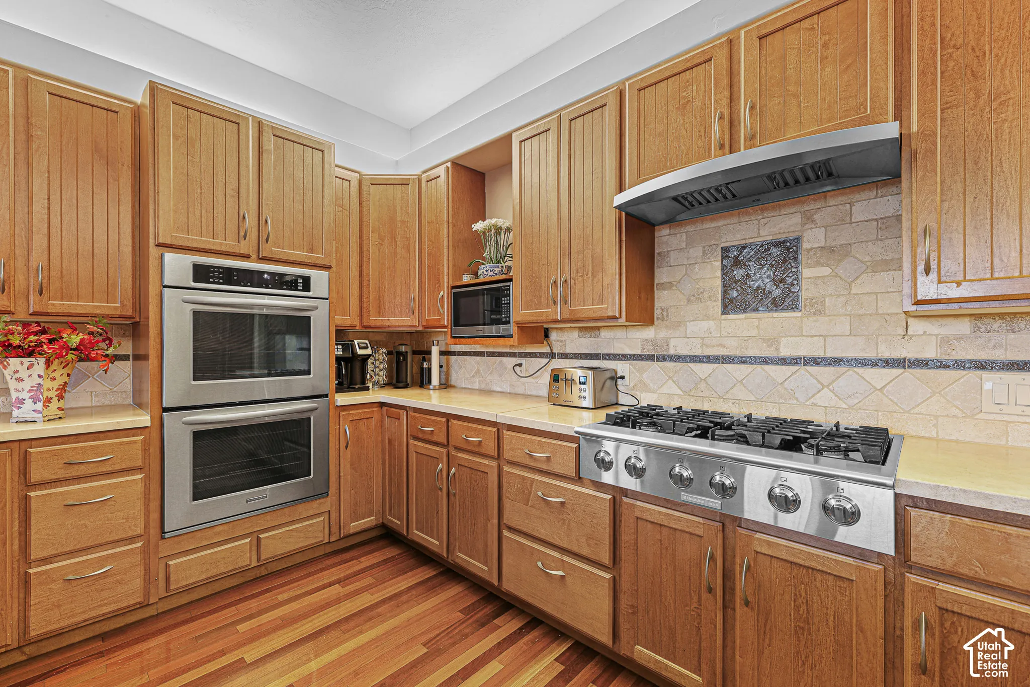 Chef's kitchen featuring warm wood cabinetry, dual wall-mounted ovens with stainless steel finish, six-burner gas cooktop, stone tile backsplash, wood-finish flooring, and stainless steel range hood