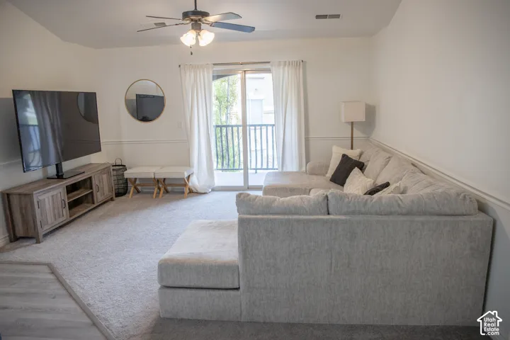 Spacious living area featuring light-toned carpeting, a ceiling fan, a sliding glass door with balcony access, and decorative wall molding