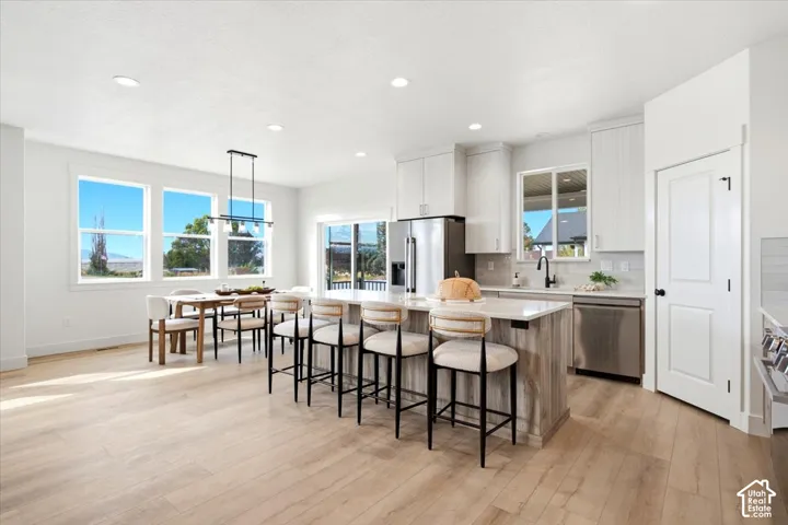 Open-concept living area with light wood-finish flooring, a kitchen island featuring a light countertop, stainless steel appliances, and multiple windows