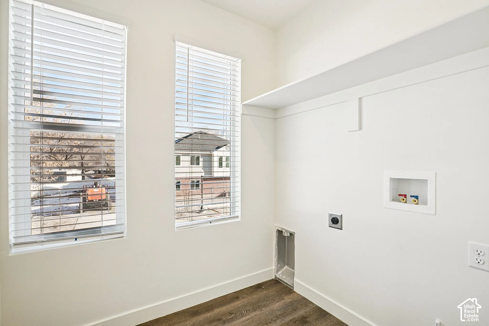 White walls, wood-finish flooring, two windows with blinds, built-in shelf, and appliance hookups