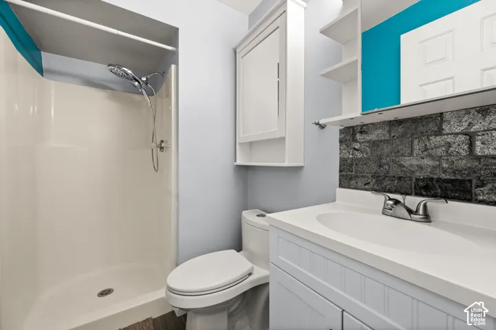 White shower enclosure with a chrome shower head and handle, white toilet, white vanity with a single basin sink, chrome faucet, and dark stone-look tile backsplash, and a white medicine cabinet with shelving