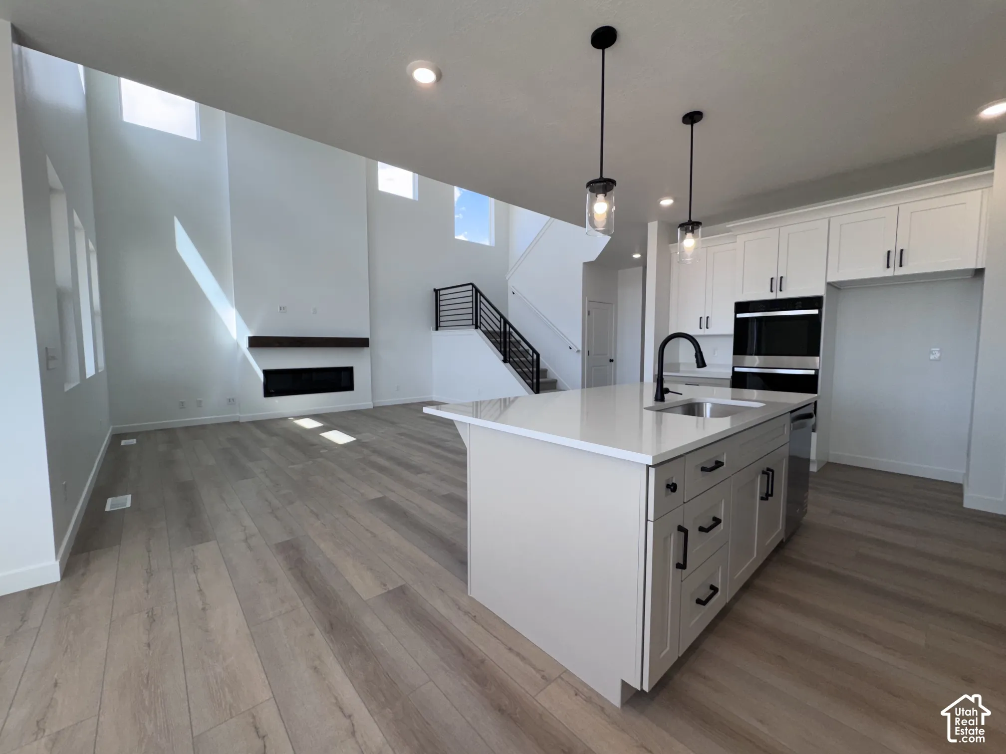 Open-concept living area featuring a kitchen island with sink, integrated double ovens, wood-finish flooring, and a fireplace with a wood-beam mantel