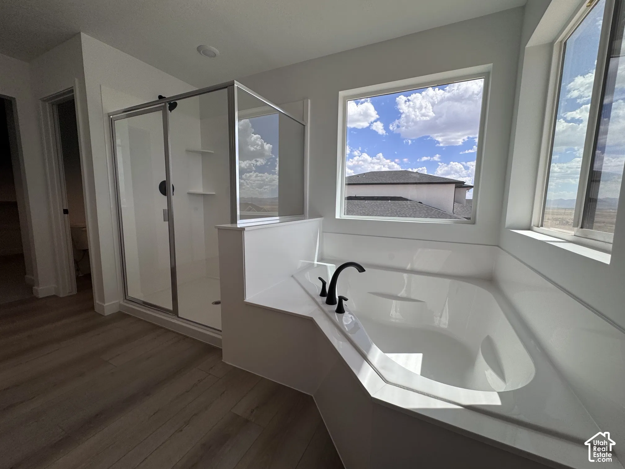 Soaking tub with matte black fixtures, glass-enclosed shower, wood-finish flooring, and multiple windows