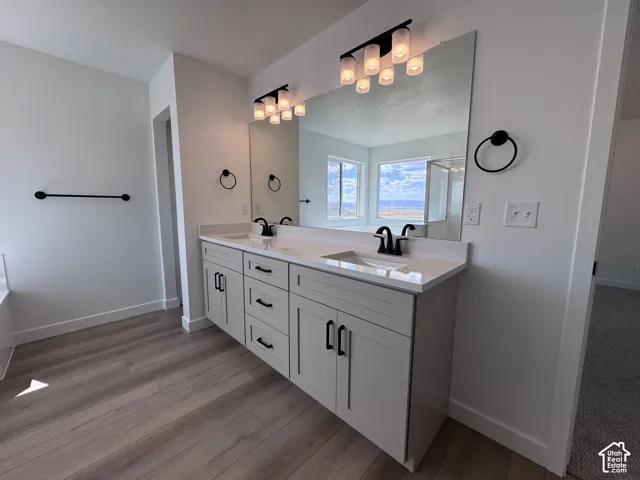 Dual vanity with white countertop and integrated sinks, matte black fixtures, wood-finish flooring, and wall-mounted lighting