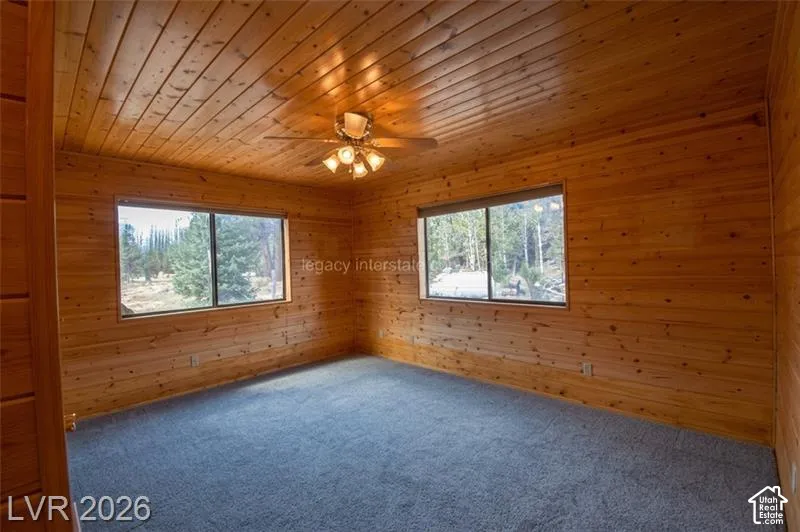 Wood paneling throughout, ceiling fan with integrated lighting, two windows with outdoor scenery, and plush carpet flooring