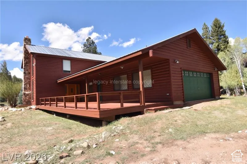 Log-style siding with a metal roof, an expansive deck with wood railing, a stone chimney, and an integrated garage with a green door