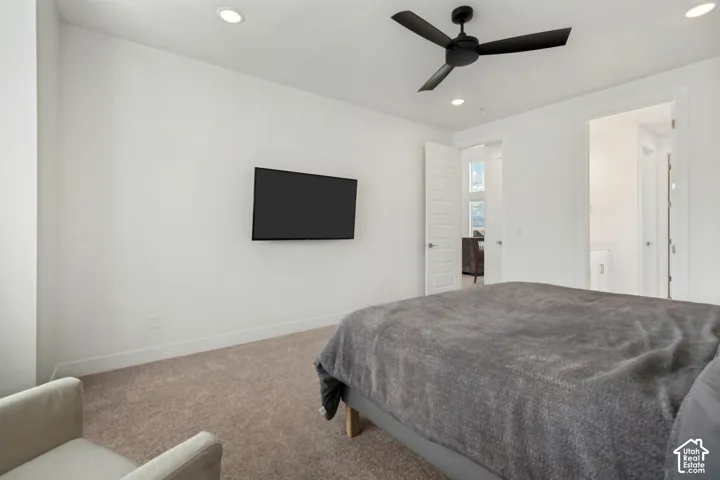 Recessed lighting illuminates a room featuring a ceiling fan with matte black blades, a mounted flat-screen, and neutral carpeting