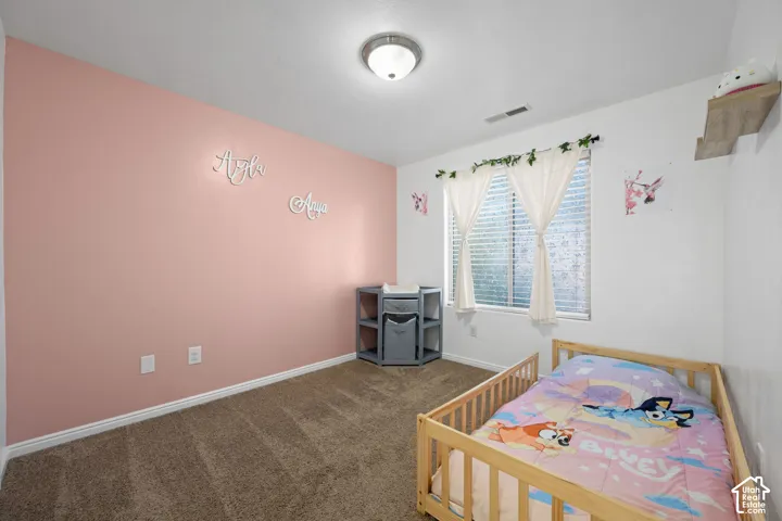 Carpeted room with a ceiling-mounted light fixture, a window featuring blinds and sheer curtains, and a built-in wall shelf