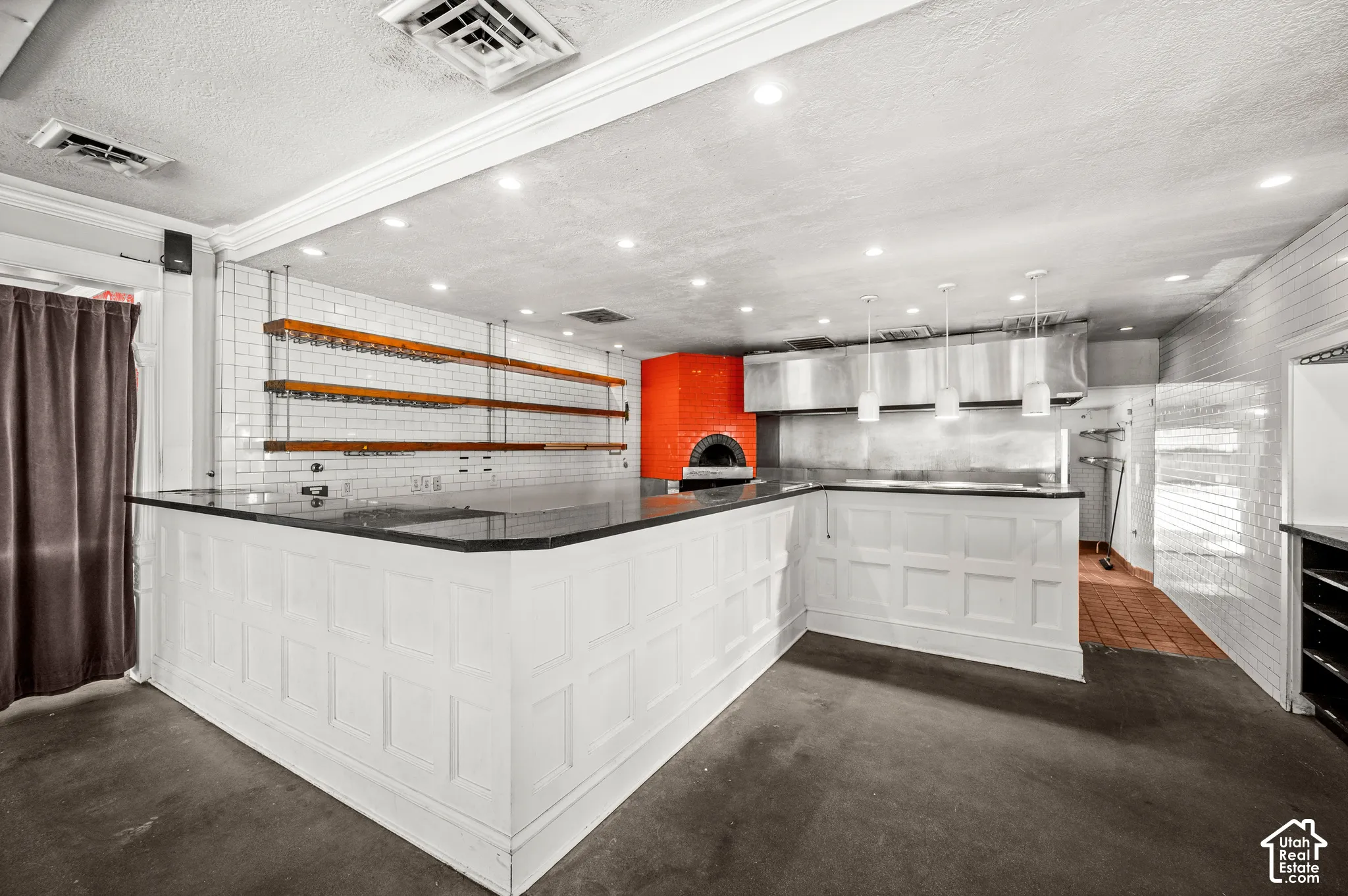 Commercial kitchen featuring white subway tile, a built-in pizza oven, stainless steel range hood, white paneled bar, and pendant lighting