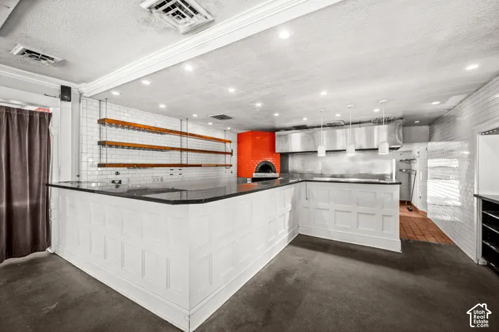Commercial kitchen featuring white subway tile, a built-in pizza oven, stainless steel range hood, white paneled bar, and pendant lighting