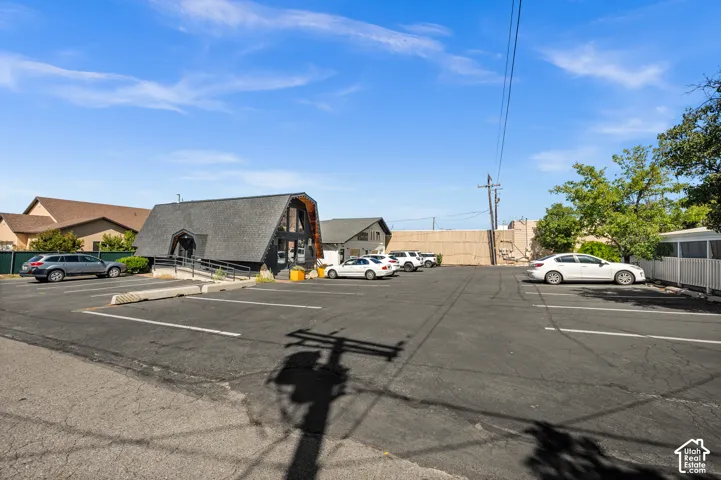 Paved parking lot with marked spaces, A-frame style building with dark siding, front entry ramp, mature trees, and perimeter fencing