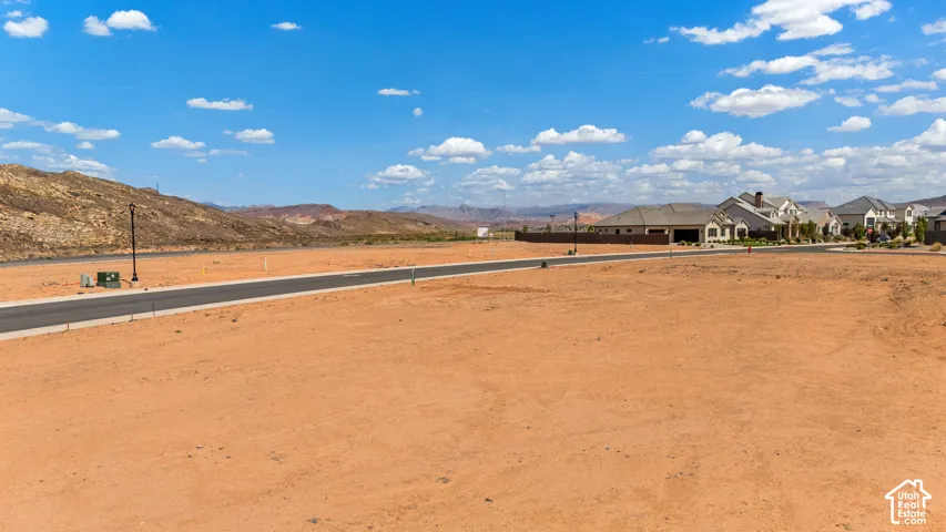 Expansive undeveloped land featuring a paved road, mountainous backdrop, and established neighboring residences