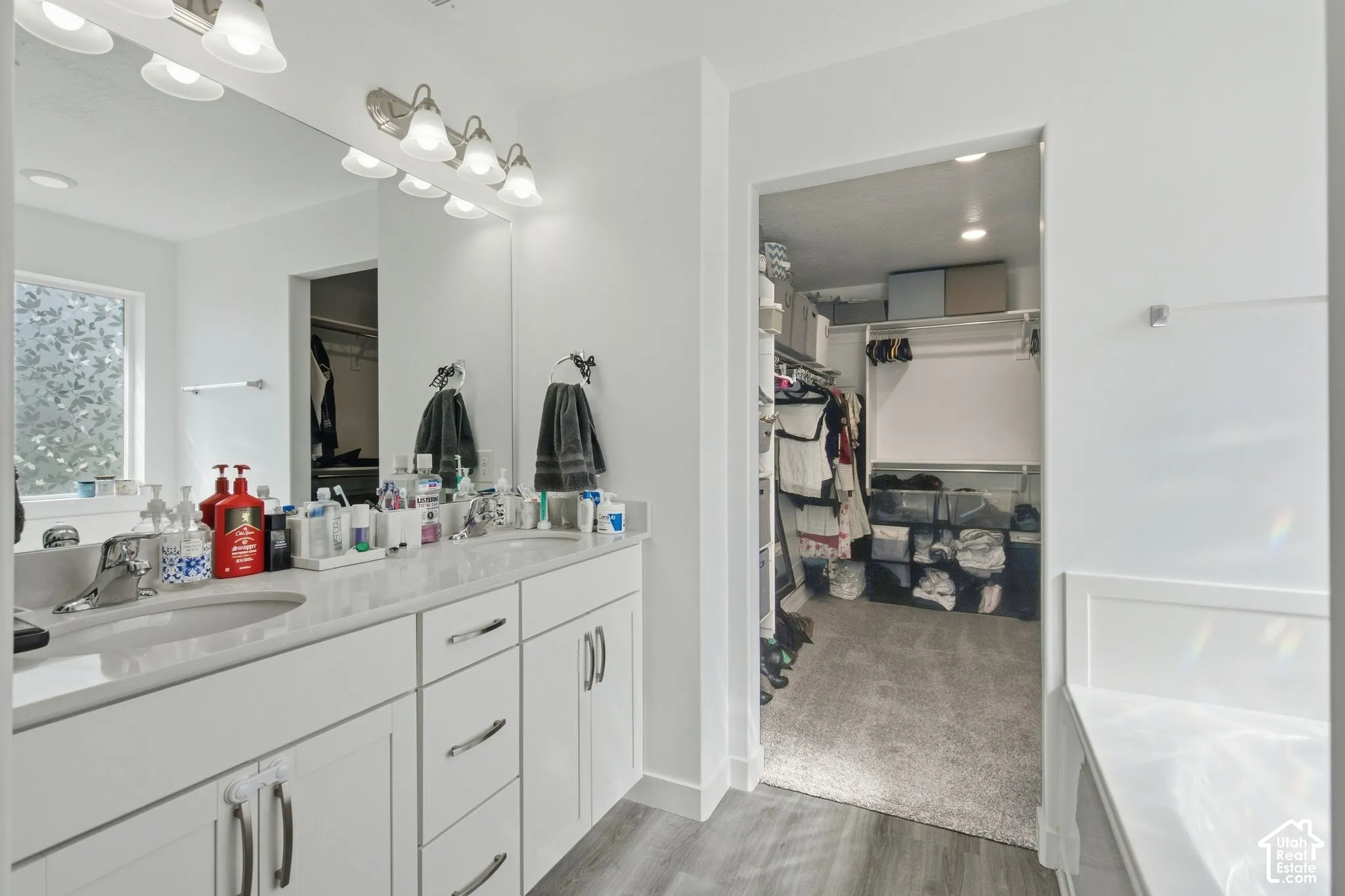 Double vanity with undermount sinks, wall-mounted light fixture, large mirror, and wood-finish flooring