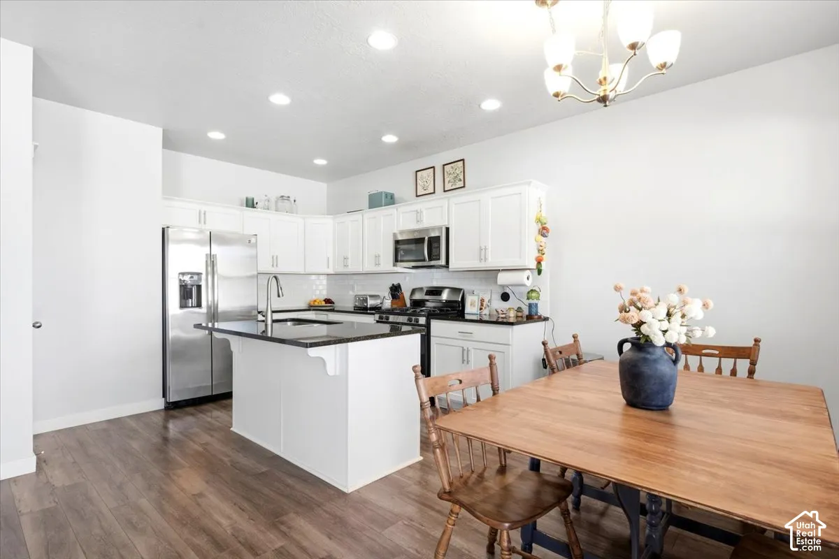 Kitchen featuring white cabinetry, dark countertops, stainless steel appliances, a central island with a sink, and wood-finish flooring