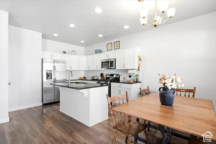 Kitchen featuring white cabinetry, dark countertops, stainless steel appliances, a central island with a sink, and wood-finish flooring