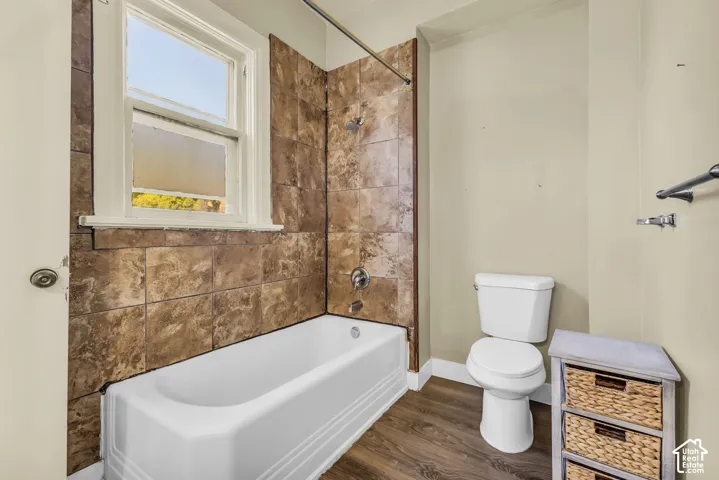 Bathtub with surrounding tile, window for natural light, toilet, and wood-finish flooring
