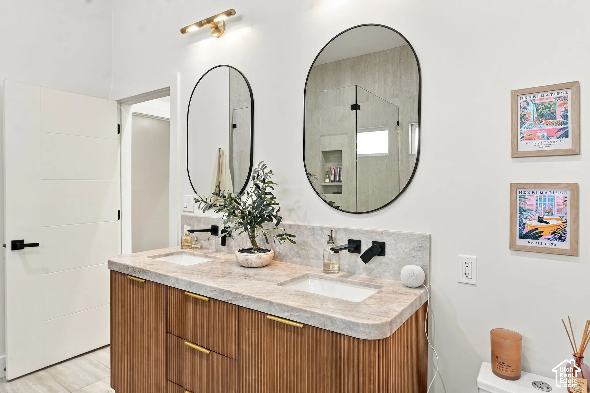 Dual oval mirrors, natural wood-finish vanity with fluted detailing, light-toned countertop, and matte black wall-mounted faucets