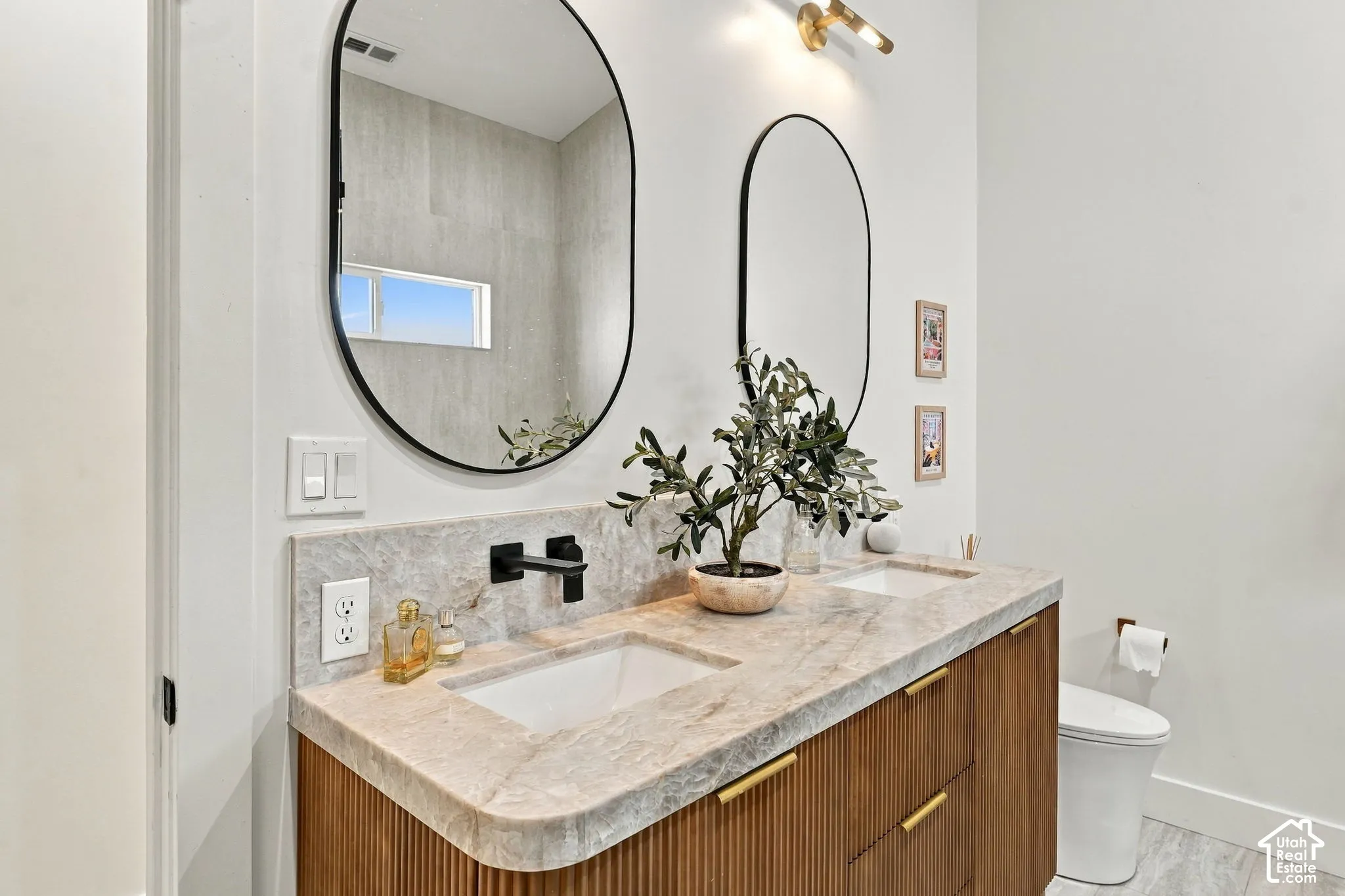 Dual wall-mounted oval mirrors, stone countertop with integrated sinks, wood-finish vanity with fluted detailing, matte black wall-mounted faucets, and gold-finish hardware