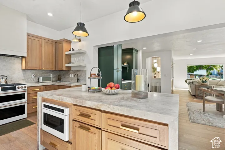 Kitchen featuring an oversized island with an integrated microwave, light wood-finish cabinetry, a white range with double ovens, and a natural stone backsplash and countertops
