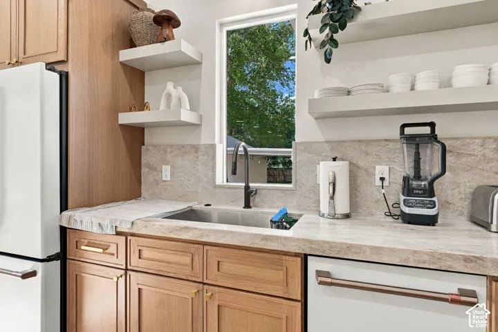 White refrigerator, wood-finish cabinetry, stainless steel sink with matte black faucet, light-toned stone countertops, and floating open shelving