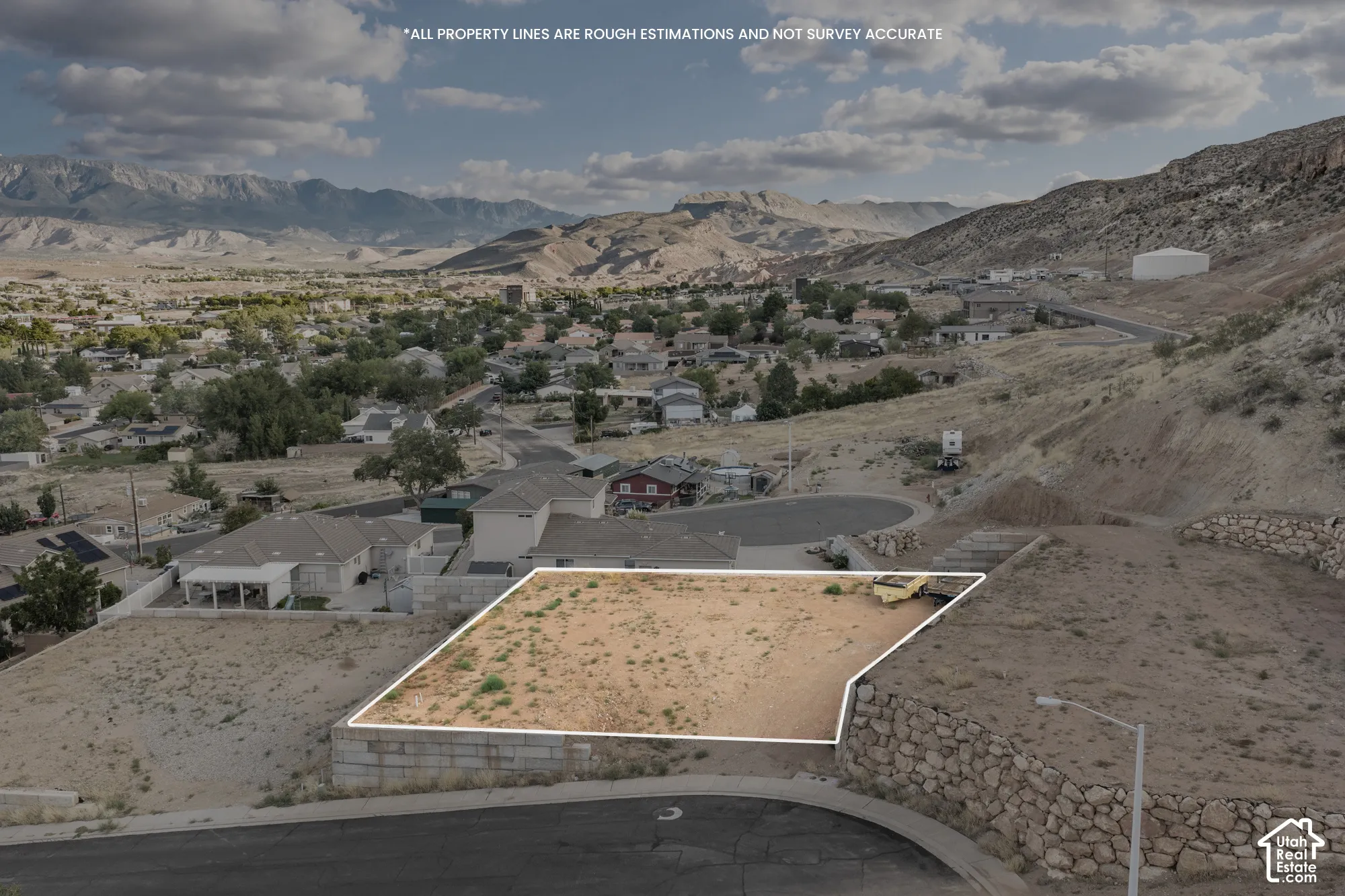 Desert landscape with a defined lot, surrounding mountainous terrain, and residential structures in the background