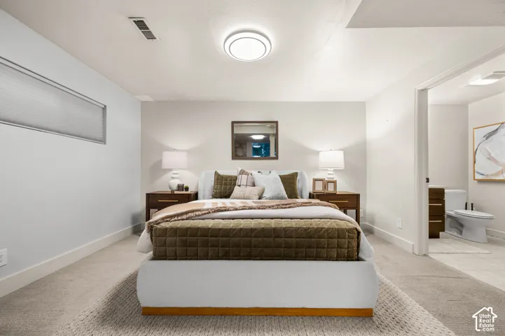 Recessed window with horizontal blinds, carpeted flooring, built-in ceiling light fixture, wood-finish nightstands, and an adjacent bathroom with tiled flooring and a toilet