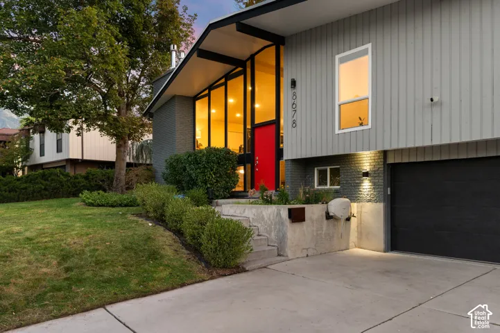 Mid-century modern exterior featuring vertical siding, extensive floor-to-ceiling windows, a vibrant red entry door, a dark garage door, and a concrete driveway