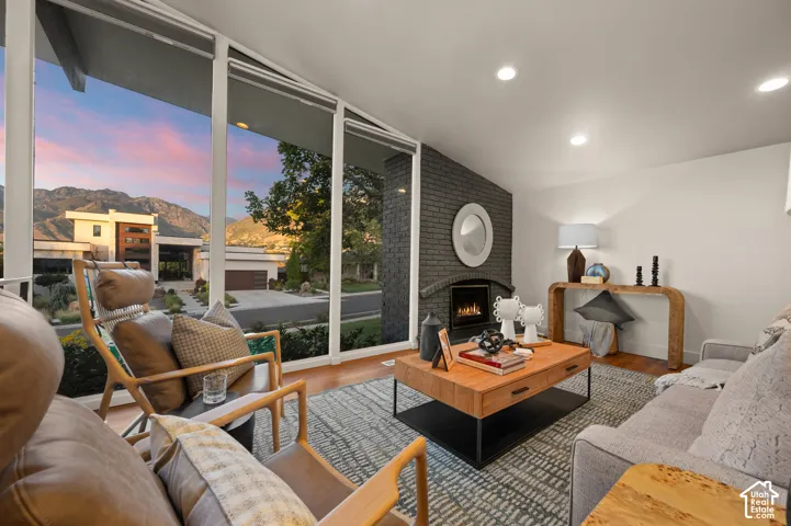 Expansive window wall, gas fireplace with brick surround, wood-finish flooring, recessed lighting, and white walls