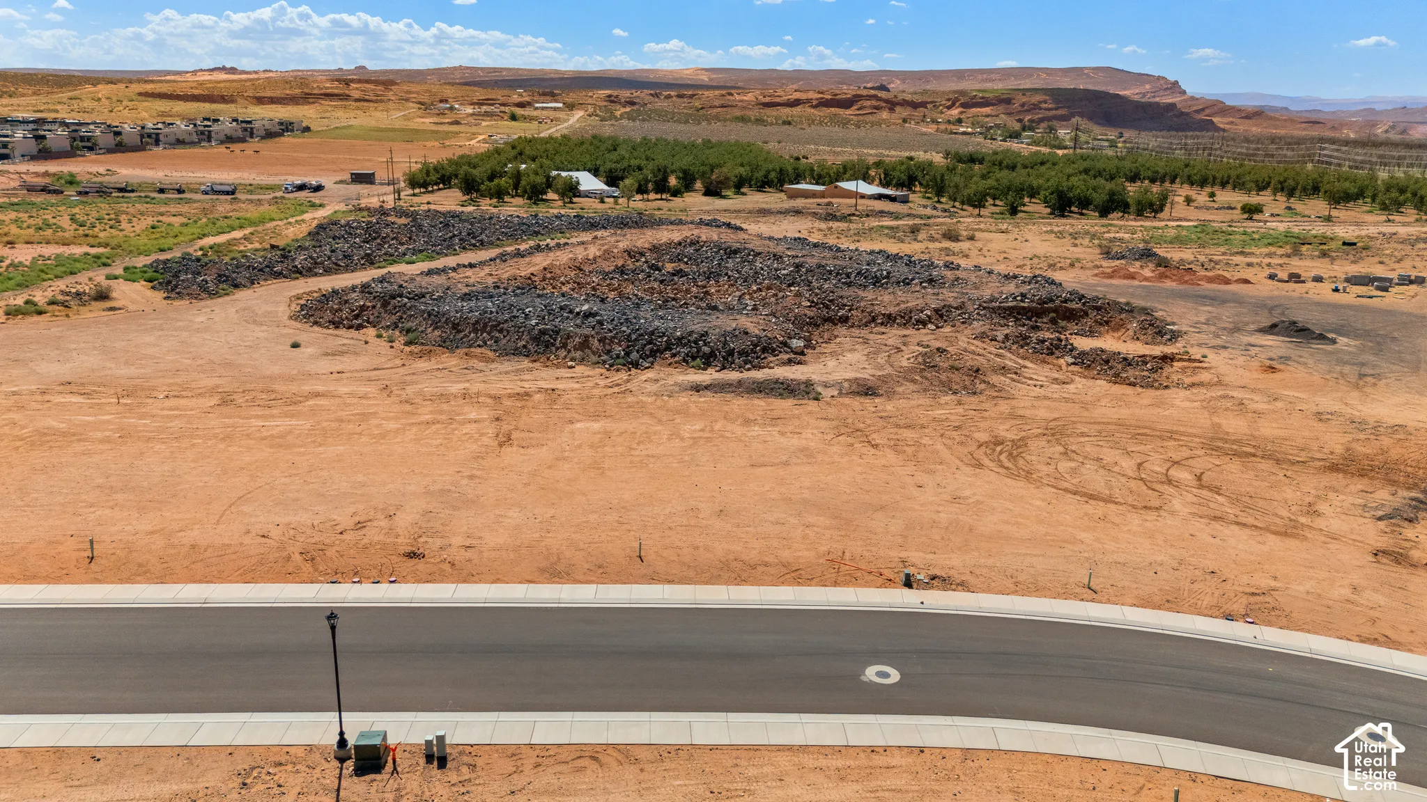 Paved street with concrete curbing, barren land with scattered rock formations, distant green foliage, and natural rock formations on the horizon
