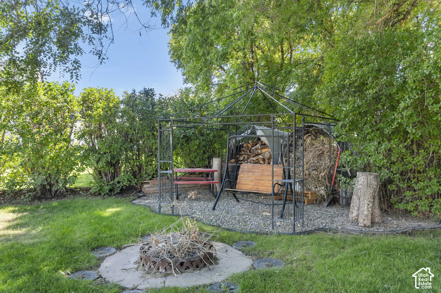 Manicured lawn, mature trees, fire pit with concrete surround, gravel patio, and metal gazebo structure