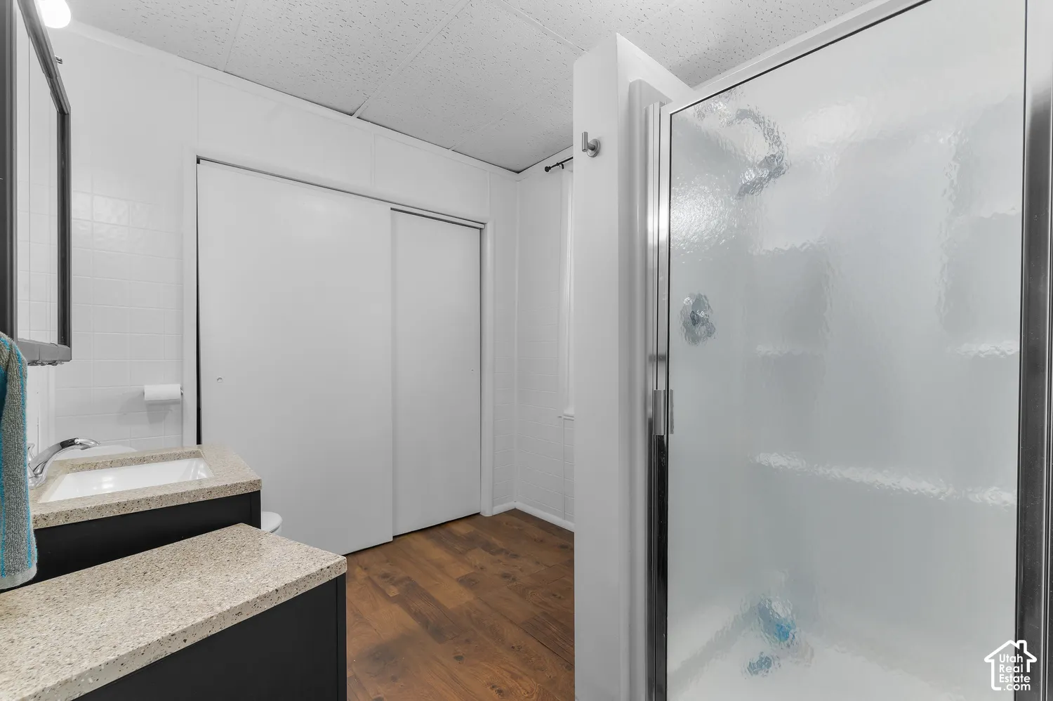 Walk-in shower with textured glass enclosure, dual vanity with undermount sinks and light-toned countertops, wood-finish flooring, and white paneling