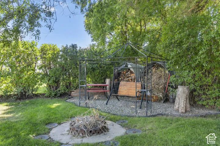 Manicured lawn, mature trees, fire pit with concrete surround, gravel patio, and metal gazebo structure