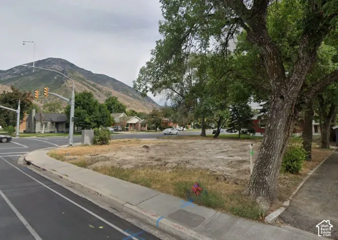 Vacant lot with mature trees, concrete sidewalk, and mountain backdrop