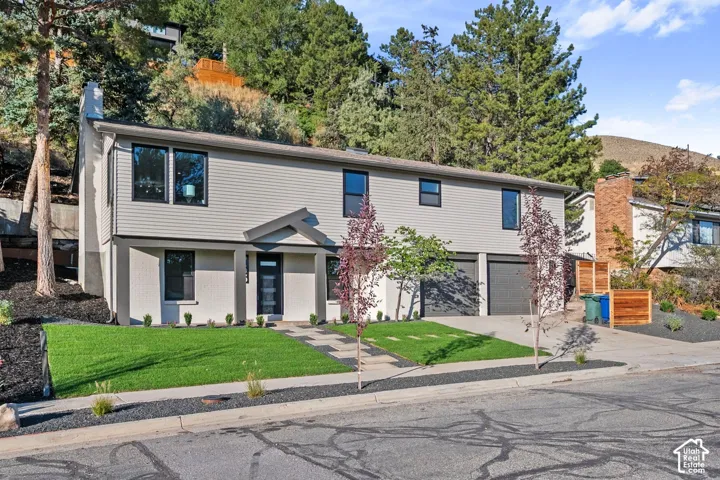 Contemporary exterior with light gray siding and stucco, featuring a prominent front entrance with a dark door, multiple dark-framed windows, two integrated garage doors, and a manicured lawn