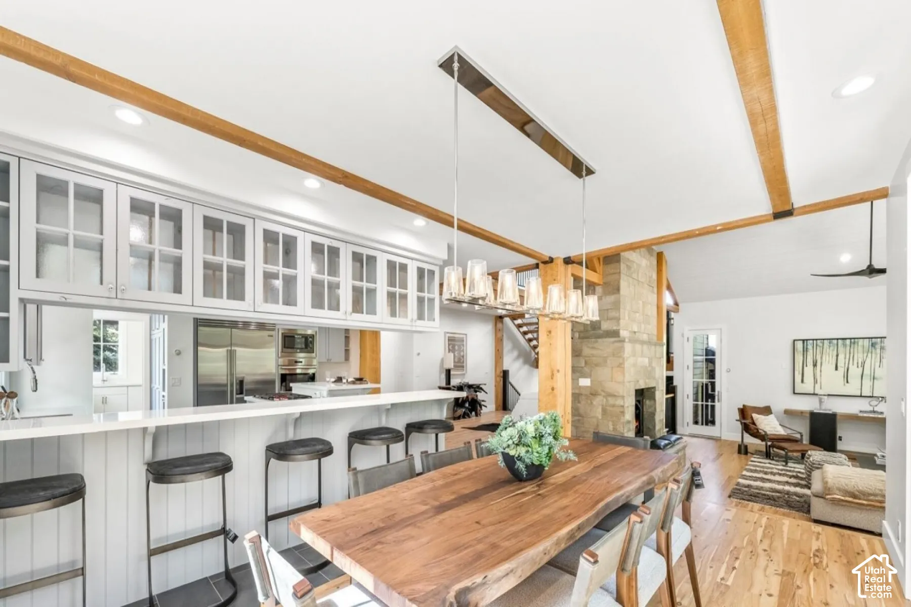 Exposed wood beams, stone-clad fireplace, wood-finish flooring, glass-front upper cabinetry, and a substantial wood dining table
