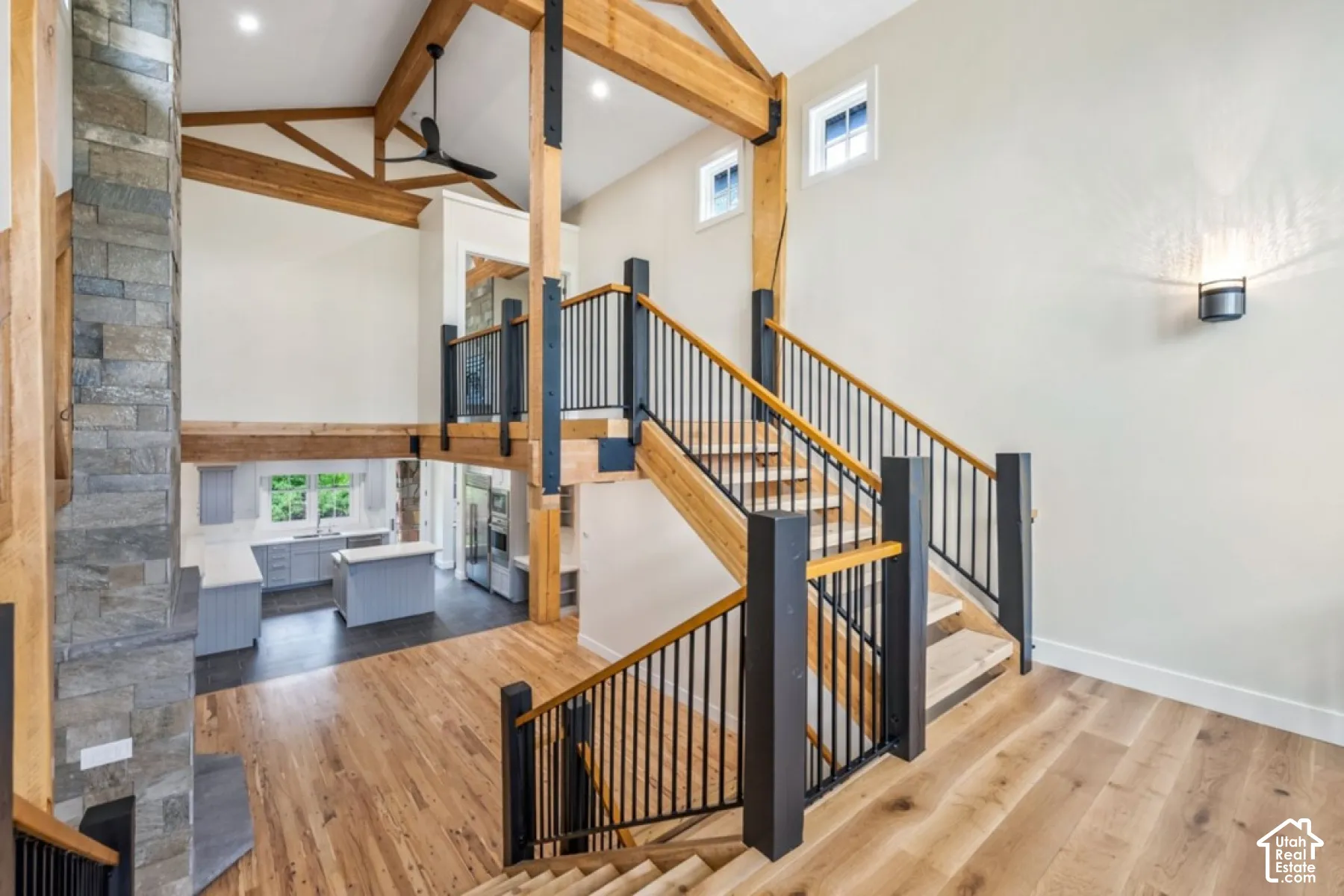 Vaulted ceilings with exposed wood beams, hardwood flooring, stone-clad pillar, open-tread staircase with black metal balusters, and a kitchen featuring light gray cabinetry
