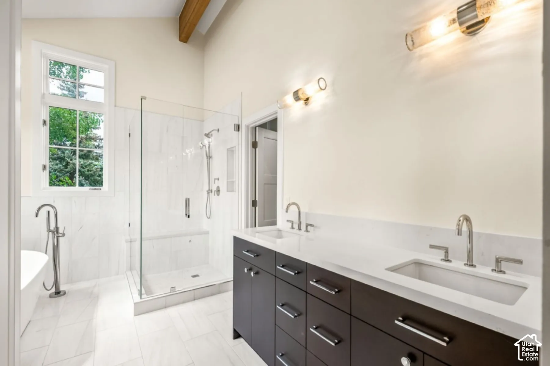 Vaulted ceiling with exposed wood beam, dual vanity with undermount sinks, glass-enclosed shower, freestanding bathtub, and large window