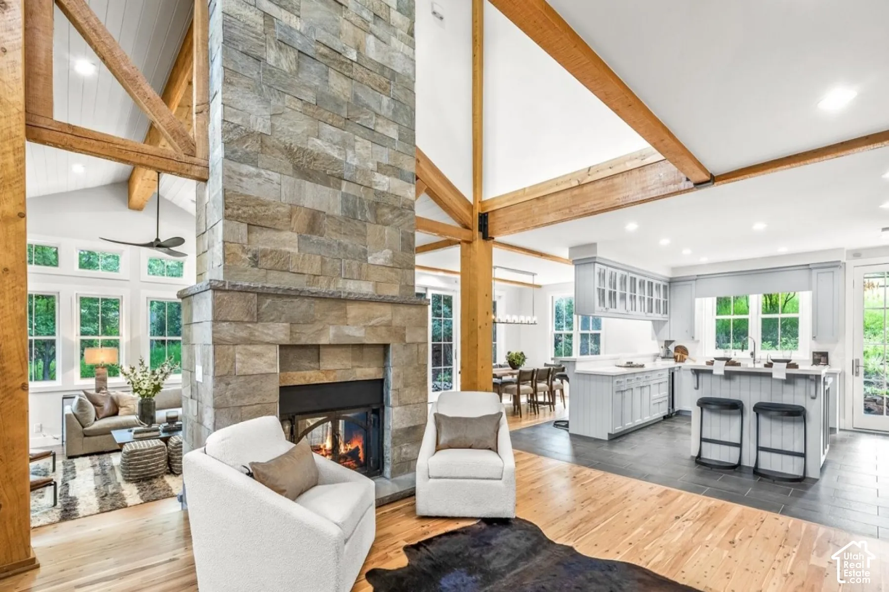 Vaulted ceilings with exposed beams, stone-faced fireplace with hearth, a kitchen island with seating, light gray cabinetry, and wood-finish flooring