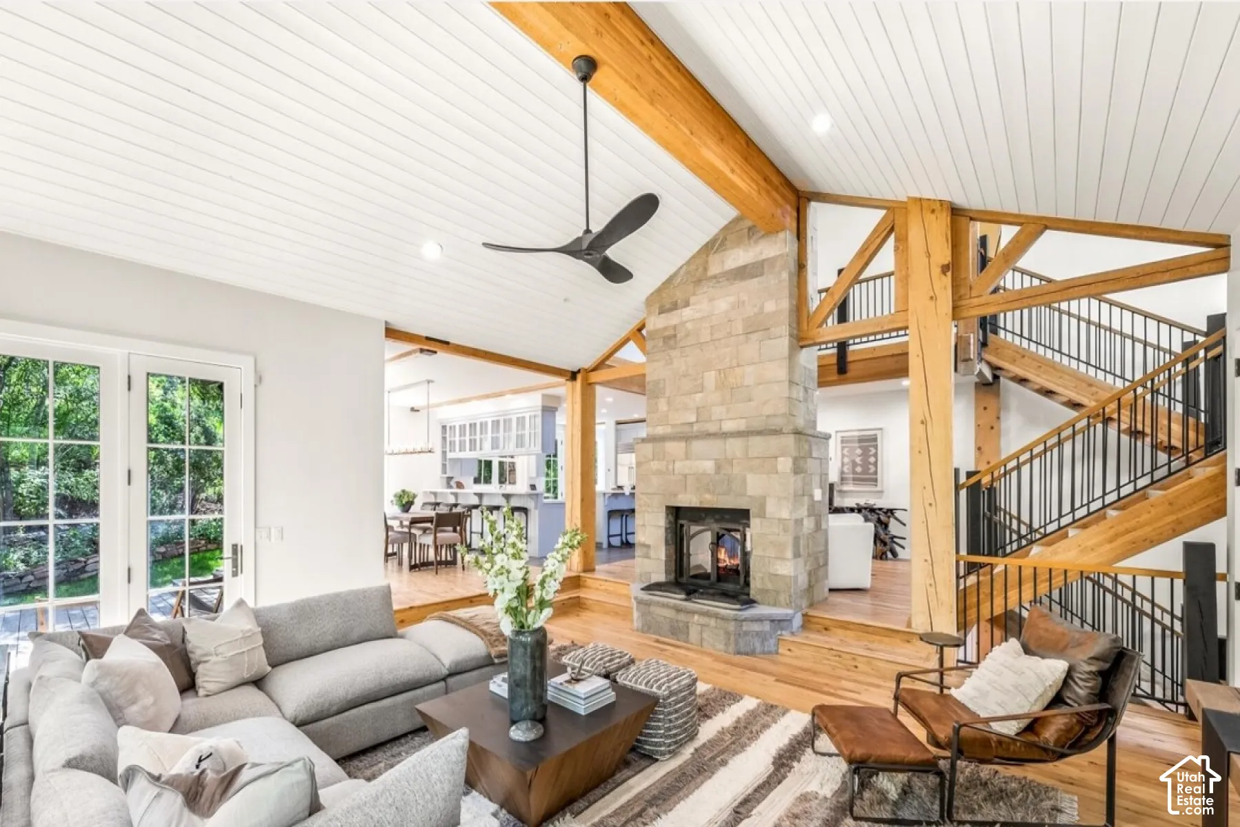 Vaulted white plank ceiling, exposed wood beams, stone-clad fireplace, hardwood flooring, and French doors