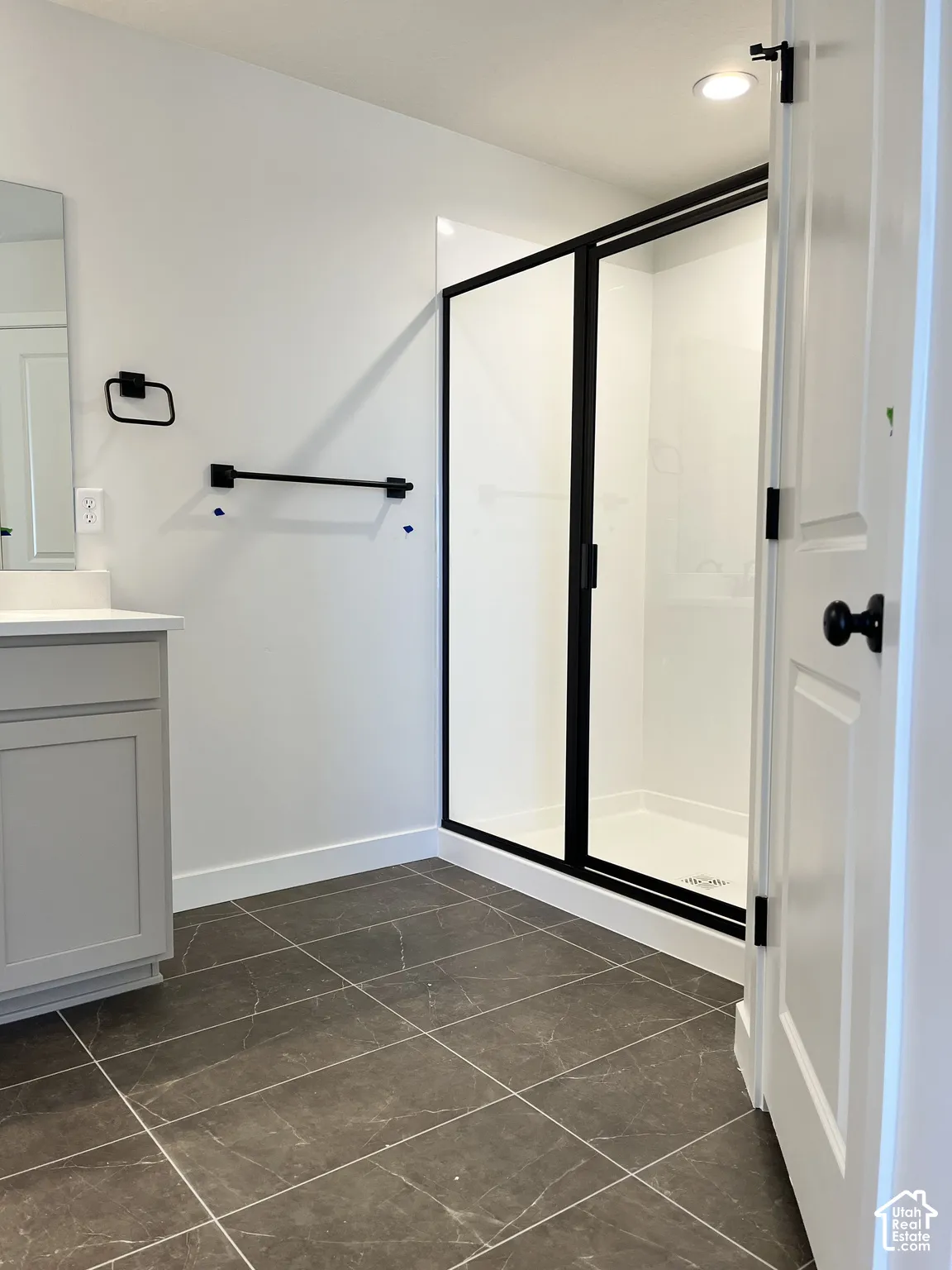 Gray tile flooring, modern vanity with white countertop, black-framed glass shower enclosure, white shower surround, and recessed lighting