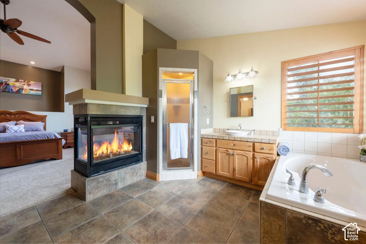 Three-sided fireplace, expansive soaking tub with deck, wood-finish vanity with vessel sink, glass-enclosed shower, and wood plantation shutters