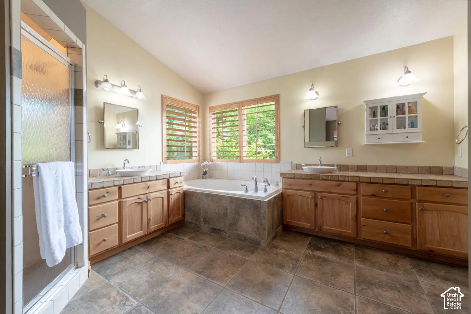 Dual wood-finish vanities with vessel sinks, a built-in soaking tub, tile flooring, and a glass-enclosed shower