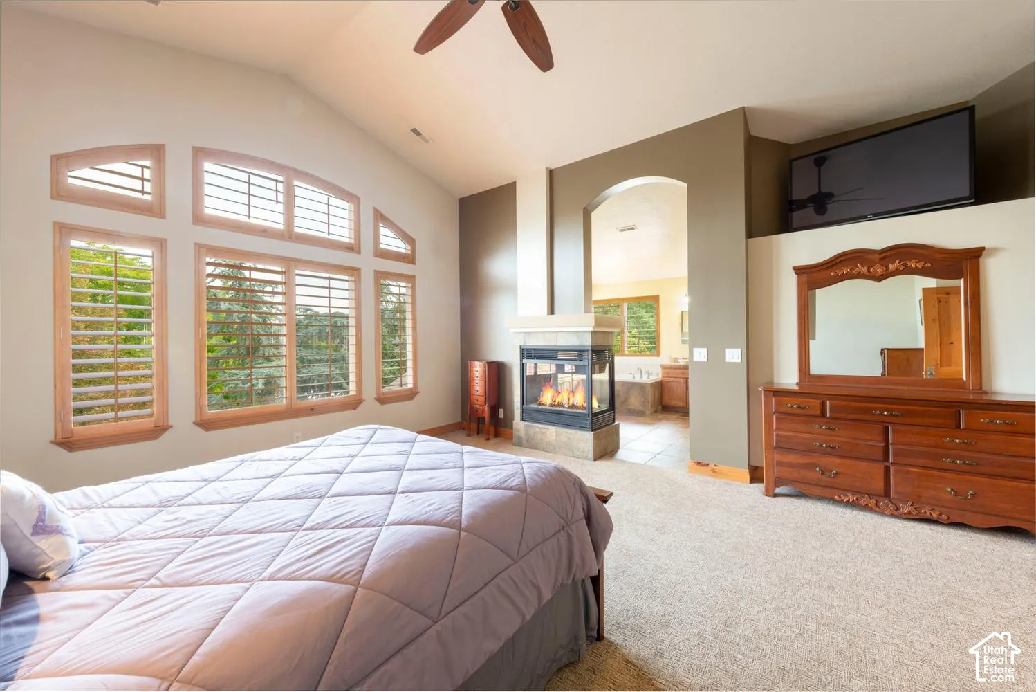 Vaulted ceiling with an installed ceiling fan, an arched room opening, a three-sided fireplace with a stone surround, wood-framed windows, and carpet flooring